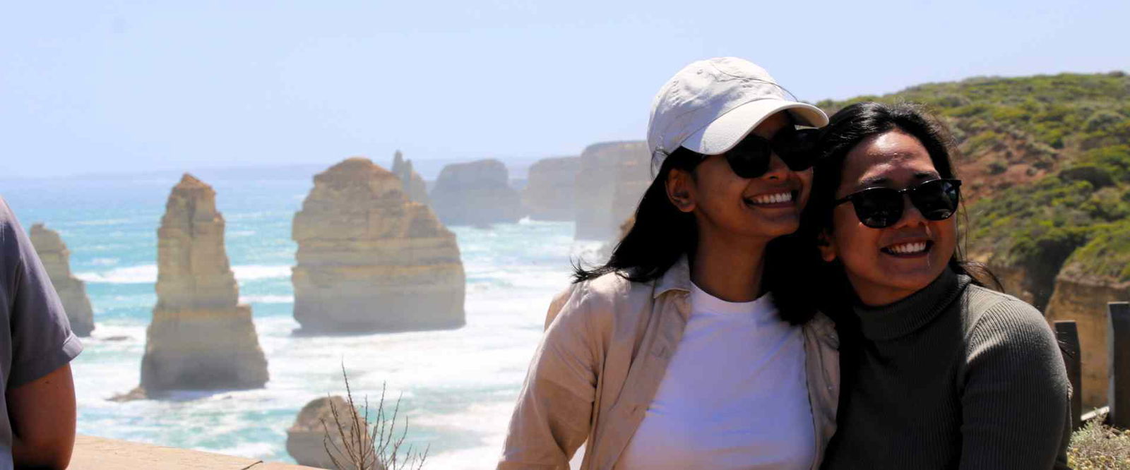 Two women smiling for a picture standing on lookout infront of the 12 apostles, on the Great Ocean Road
