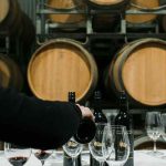 Person serving wine on an empty table set for a wine tasting in a winery with many wine barrles in the background
